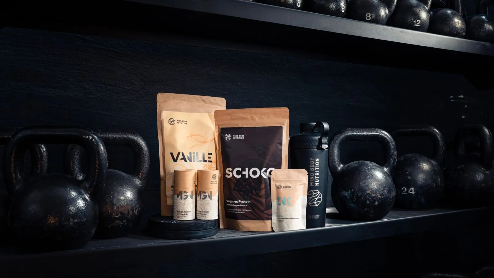 Mind Body Nutrition supplement pouches and a shaker bottle displayed on a shelf among gym kettlebells