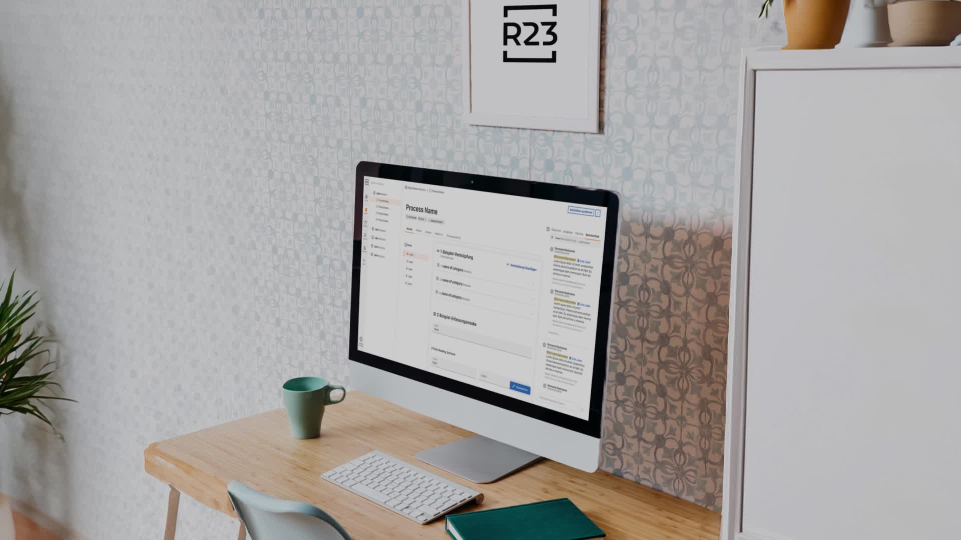 An iMac on a desk displaying the R23 process-builder interface, with a framed R23 logo on the wall behind it
